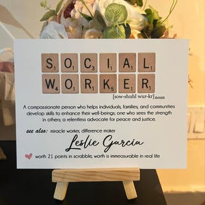May include: A white card with the word "SOCIAL WORKER" spelled out in Scrabble tiles. The definition of a social worker is written below the tiles. The name "Leslie Garcia" is written in cursive below the definition. A small pink heart is in the top left corner of the card. The card is sitting on a small wooden easel.