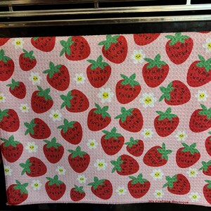 May include: A pink kitchen towel with a pattern of red strawberries, green leaves, and white daisies. Each strawberry has the words "Trump Sucks" printed on it. The towel is made of a soft material and is suitable for kitchen use.