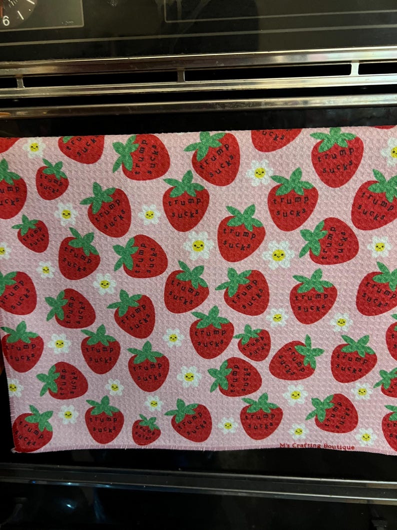 May include: A pink kitchen towel with a pattern of red strawberries, green leaves, and white daisies. Each strawberry has the words "Trump Sucks" printed on it. The towel is made of a soft material and is suitable for kitchen use.