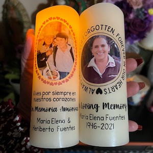 May include: Two ivory-colored memorial candles. One candle has a photo of two people and the text "Vives por siempre en nuestros corazones". The other candle has a photo of a woman and the text "Loving Memory".