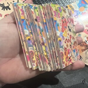 May include: A stack of colorful cards with various illustrations, including a pineapple, a car, a cat, a heart, and a snake. The cards are all different sizes and shapes, and they are arranged in a random order.