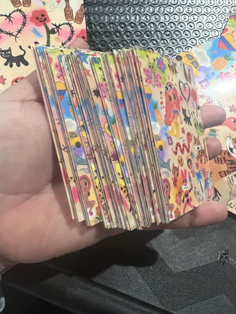 May include: A stack of colorful cards with various illustrations, including a pineapple, a car, a cat, a heart, and a snake. The cards are all different sizes and shapes, and they are arranged in a random order.