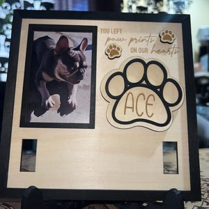 May include: A wooden pet memorial frame with a photo of a dog. The frame includes the text "YOU LEFT paw prints ON OUR hearts" and a paw print design with the name "ACE". The frame is supported by a black stand.