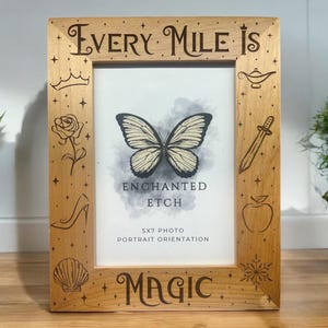 May include: A wooden picture frame with the words "Every Mile is Magic" engraved on it. The frame has a 5x7 photo opening and is decorated with various Disney-themed images, including a crown, a rose, a butterfly, a genie lamp, a sword, an apple, a slipper, a seashell, and a snowflake.