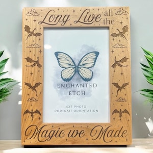 Puede incluir: Un marco de fotos de madera con un acabado marrón claro. El marco tiene un diseño grabado de dragones, nubes y estrellas. El texto "Long Live all the Magic we Made" está grabado en la parte inferior del marco. El marco está diseñado para sostener una foto de 13x18 cm en orientación vertical.