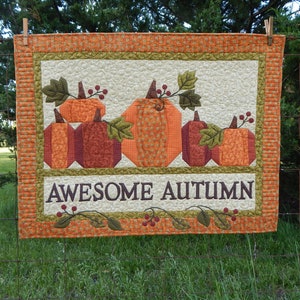 May include: A quilted wall hanging with a fall theme. It features five orange and brown pumpkins with green leaves and red berries. The text "AWESOME AUTUMN" is stitched in brown thread on the bottom.