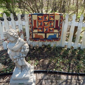 May include: A colorful patchwork quilt with a tree and cat design hangs on a white picket fence. The quilt is made of various fabrics in shades of brown, blue, red, and yellow. A stone statue of a cherub holding a bouquet of flowers sits in the foreground.