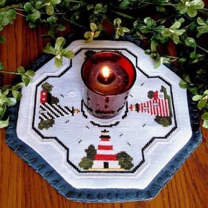May include: An octagonal white fabric mat featuring cross-stitched lighthouse designs in red, white, and black, with a dark blue stitched border. A dark red glass candle holder with a lit candle sits in the center of the mat.