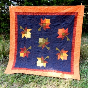 May include: A quilt with a navy blue background and a patchwork design of orange, yellow, and green maple leaves. The quilt is edged with a wide orange border.
