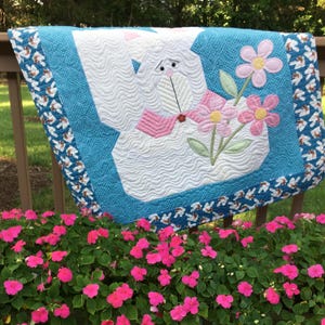 May include: A white quilted bunny with pink bow and flowers, on a blue background with a white and brown border. The bunny is holding a bouquet of pink flowers with green leaves. The quilt is hanging on a wooden railing with a green background.