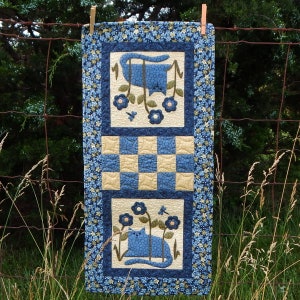 Könnte beinhalten: Ein blauer und weißer gesteppter Tischläufer mit einem karierten Muster und zwei blauen Katzen mit floralen Akzenten. Der Läufer hängt an einem Zaun.