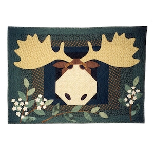 May include: A green and brown patchwork quilt with a moose head design. The moose has large antlers and a brown nose. The quilt is surrounded by a border of green leaves and white flowers.