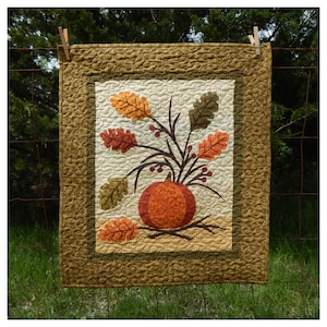 May include: A quilted wall hanging with a brown, orange, and green pumpkin and leaves design. The background is a cream color with a brown and green border.