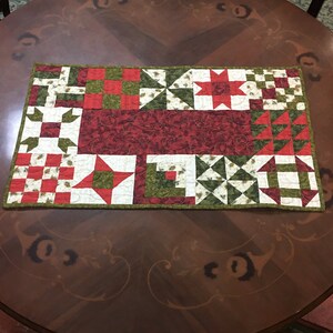 Peut inclure: Un chemin de table matelassé avec un motif patchwork composé de carrés, d'étoiles et de triangles rouges, verts et blancs. Le chemin de table est en tissu de coton et a un style rustique, de ferme.