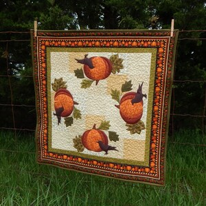 May include: A quilted wall hanging with an autumn theme. It features four orange pumpkins with black birds and green leaves on a white background. The border is brown, orange, and green with a repeating pattern of small pumpkins.