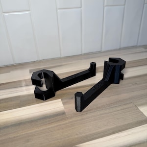 Könnte beinhalten: Zwei schwarze, 3D-gedruckte Werkzeuge in Form eines Schraubenschlüssels. Jedes Werkzeug hat eine sechseckige Öffnung und eine hakenartige Verlängerung. Die Werkzeuge liegen auf einer hellbraunen Oberfläche mit weißem Fliesenhintergrund. Der Text "W3D" ist sichtbar.