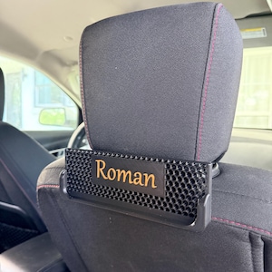 Könnte beinhalten: Schwarze Auto-Kopfstütze mit einem personalisierten Namensschild mit dem goldenen Schriftzug "Roman". Das Schild ist an einem schwarzen, perforierten Halter befestigt. Die Kopfstütze ist schwarz mit roten Nähten.