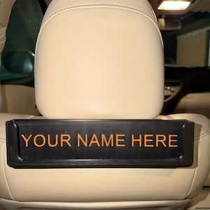 Könnte beinhalten: Schwarzes Kopfstützenzubehör für Autos mit dem Text "YOUR NAME HERE" in Orange. Das Zubehör ist rechteckig und sitzt auf einem beigefarbenen Autositz. Die Kopfstütze ist ebenfalls beige.