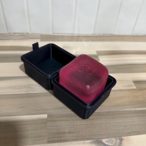 Op de afbeelding: Een open zwarte rechthoekige doos met een roze, doorschijnend, vierkant object. De doos staat op een houten oppervlak, met een witte betegelde achtergrond.