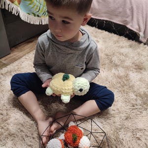 May include: A child holds a handmade crochet turtle toy with a yellow and light green shell. Several other crochet turtles in orange, white, and gray are in a black wire basket. The toys are on a fluffy, cream-colored rug.