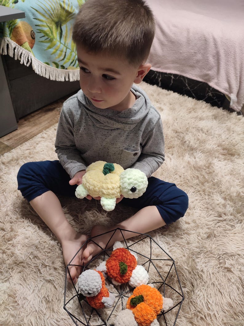 May include: A child holds a handmade crochet turtle toy with a yellow and light green shell. Several other crochet turtles in orange, white, and gray are in a black wire basket. The toys are on a fluffy, cream-colored rug.