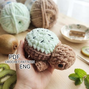 May include: A crochet turtle toy with a light green, textured shell resembling a kiwi fruit, featuring small black embroidered seeds. The head and limbs are made of brown yarn, and the toy includes a black plastic eye. Text reads PDF CROCHET PATTERN ENG.
