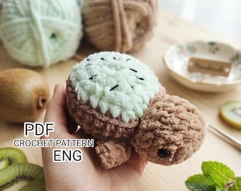 May include: A crochet turtle toy with a light green, textured shell resembling a kiwi fruit, featuring small black embroidered seeds. The head and limbs are made of brown yarn, and the toy includes a black plastic eye. Text reads PDF CROCHET PATTERN ENG.
