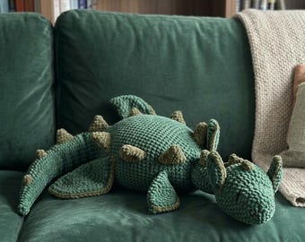 May include: A handmade, green crochet dinosaur plush toy with brown accents, resting on a green velvet sofa. The dinosaur has a textured body, detailed limbs, and a friendly expression. The plush is a soft, decorative item.