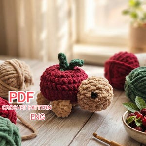 May include: A crochet turtle toy with a red apple-shaped shell featuring green leaves, a tan head with a black eye, and tan feet. The text PDF CROCHET PATTERN ENG is visible on the left. Several balls of yarn and a bowl of berries are nearby.
