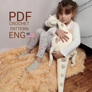 Puede incluir: Un peluche de dinosaurio de ganchillo de color crema con detalles en beige. El juguete lo sostiene una persona. La imagen también incluye el texto "PDF CROCHET PATTERN ENG" con una bandera americana.