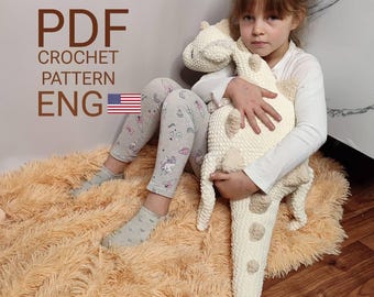 May include: A cream-colored, crocheted dinosaur plush toy with tan accents. The toy is held by a person. The image also includes the text "PDF CROCHET PATTERN ENG" with an American flag.