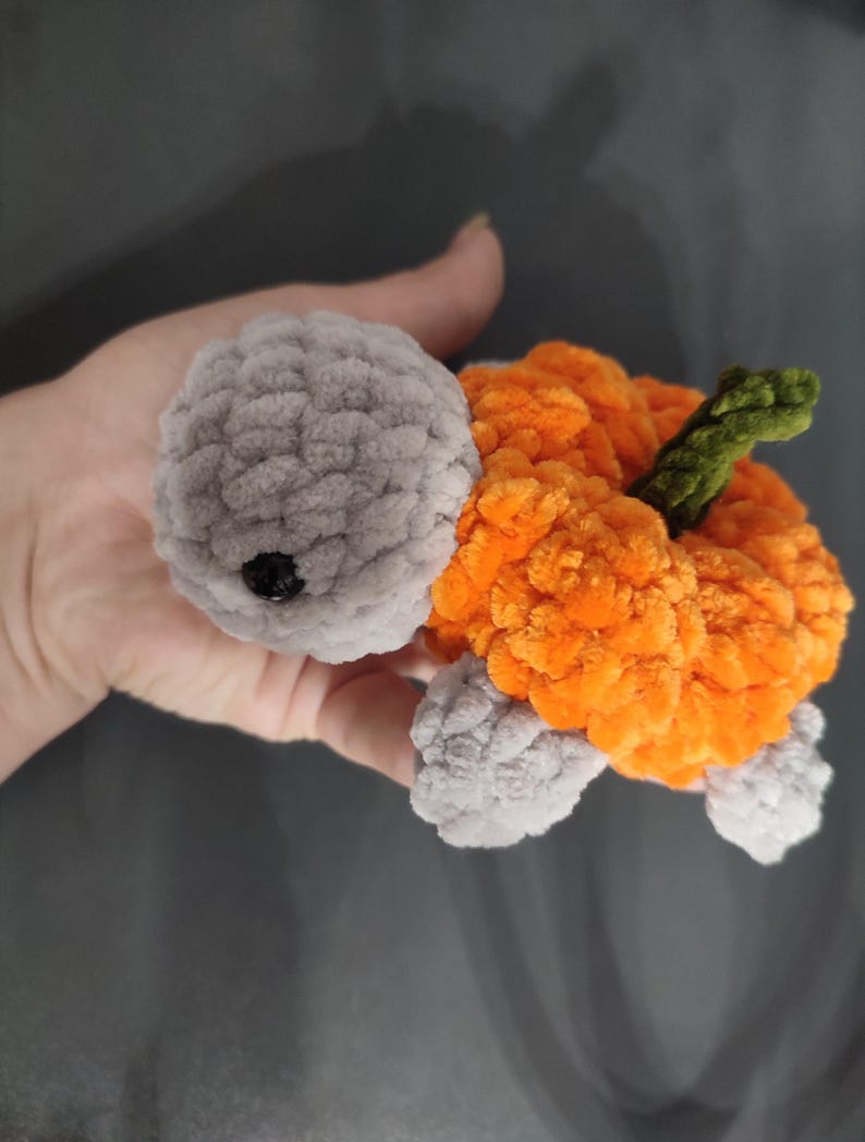 May include: A handmade crochet turtle with a gray head and feet, and an orange body resembling a pumpkin. The turtle has a green stem and a black eye. The plush toy is made of soft, textured yarn.