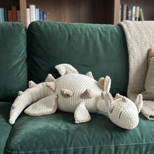 Puede incluir: Un cojín con forma de dinosaurio tejido a ganchillo en crema y marrón, apoyado sobre un sofá de terciopelo verde oscuro. El peluche presenta detalles texturizados y está rodeado por una manta y cojines de tonos neutros.