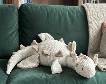 May include: A crocheted dinosaur-shaped pillow in cream and tan, resting on a dark green velvet couch. The plush toy features textured details and is surrounded by a neutral-toned blanket and pillows, creating a cozy aesthetic.