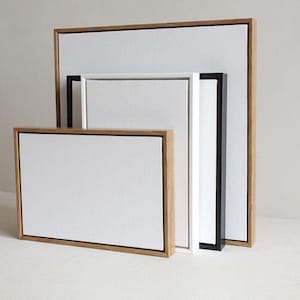 May include: Several rectangular canvas art pieces with white surfaces and different frame styles. The frames are natural wood, black, and white, offering a minimalist look. The sizes vary, creating a layered display.