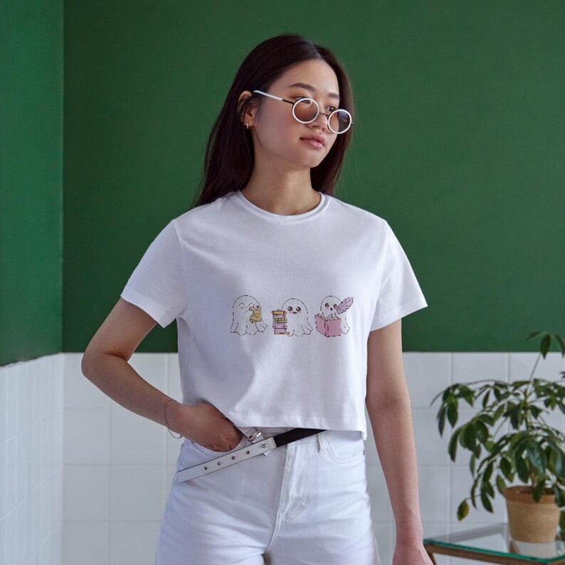 K&ouml;nnte beinhalten: Wei&szlig;es Crop-Top mit einer skurrilen Illustration von drei Cartoon-Geistern. Die Geister sind mit verschiedenen Gegenst&auml;nden dargestellt, darunter ein Buch und eine Feder. Das Shirt wird mit wei&szlig;en Shorts und einem silbernen G&uuml;rtel kombiniert.