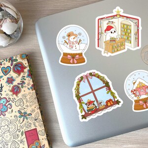 May include: A collection of colorful Christmas-themed stickers on a laptop. Designs include snow globes with snowmen and houses, a window scene, and a ghost reading in a book. A diary with floral patterns is also visible.