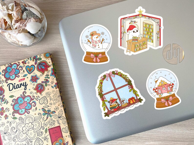 May include: A collection of colorful Christmas-themed stickers on a laptop. Designs include snow globes with snowmen and houses, a window scene, and a ghost reading in a book. A diary with floral patterns is also visible.