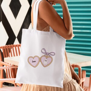 K&ouml;nnte beinhalten: Wei&szlig;e Tragetasche mit einem herzf&ouml;rmigen Design mit dem Wort "boo" und einer Geisterillustration. Das Design verwendet Pastellfarben mit einer lavendelfarbenen Schleife. Die Tasche hat zwei wei&szlig;e Griffe.