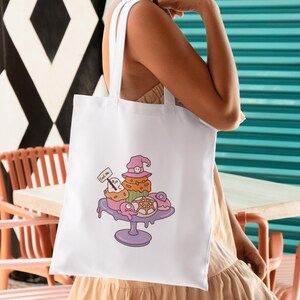 May include: White tote bag featuring a whimsical illustration of Halloween-themed cupcakes and donuts on a purple cake stand. The cupcakes are decorated with witch hats and ghosts. A small sign reads "Eat me".