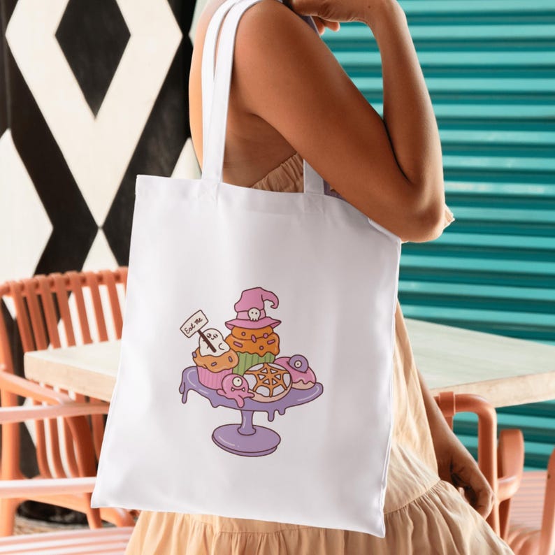 May include: White tote bag featuring a whimsical illustration of Halloween-themed cupcakes and donuts on a purple cake stand. The cupcakes are decorated with witch hats and ghosts. A small sign reads "Eat me".