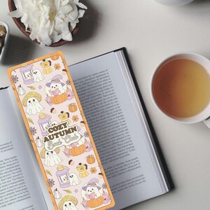 May include: A colorful bookmark with a fall theme, featuring illustrations of ghosts, pumpkins, and mushrooms. The bookmark reads "Cozy Autumn Book Club" and is placed on an open book. A cup of tea and a bowl of nuts and flowers are also visible.