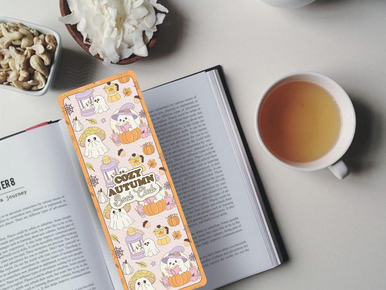 May include: A colorful bookmark with a fall theme, featuring illustrations of ghosts, pumpkins, and mushrooms. The bookmark reads "Cozy Autumn Book Club" and is placed on an open book. A cup of tea and a bowl of nuts and flowers are also visible.