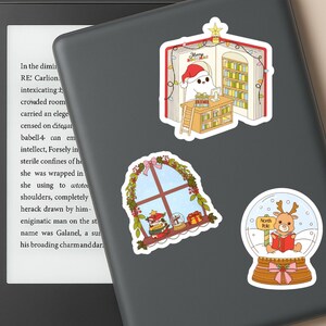 May include: A collection of Christmas-themed stickers. One sticker shows a ghost-like figure in a Santa hat in a library. Another shows a window with Christmas decorations. The last sticker shows a reindeer in a snow globe.