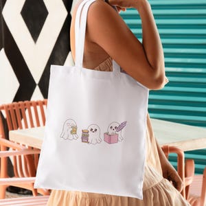 K&ouml;nnte beinhalten: Wei&szlig;e Tragetasche mit drei Cartoon-Geistern. Einer h&auml;lt Geld, ein anderer stapelt B&uuml;cher und der dritte schreibt mit einem Federkiel in ein rosa Buch. Die Tasche hat wei&szlig;e Tr&auml;ger und wird von einer Person gehalten.