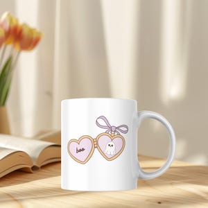 K&ouml;nnte beinhalten: Wei&szlig;e Keramiktasse mit herzf&ouml;rmigem Design, dem Wort "boo" und einer Geisterillustration. Das Design ist in Rosa-, Beige- und Lilat&ouml;nen gehalten. Die Tasse hat einen gebogenen Henkel und steht auf einer Holzoberfl&auml;che.