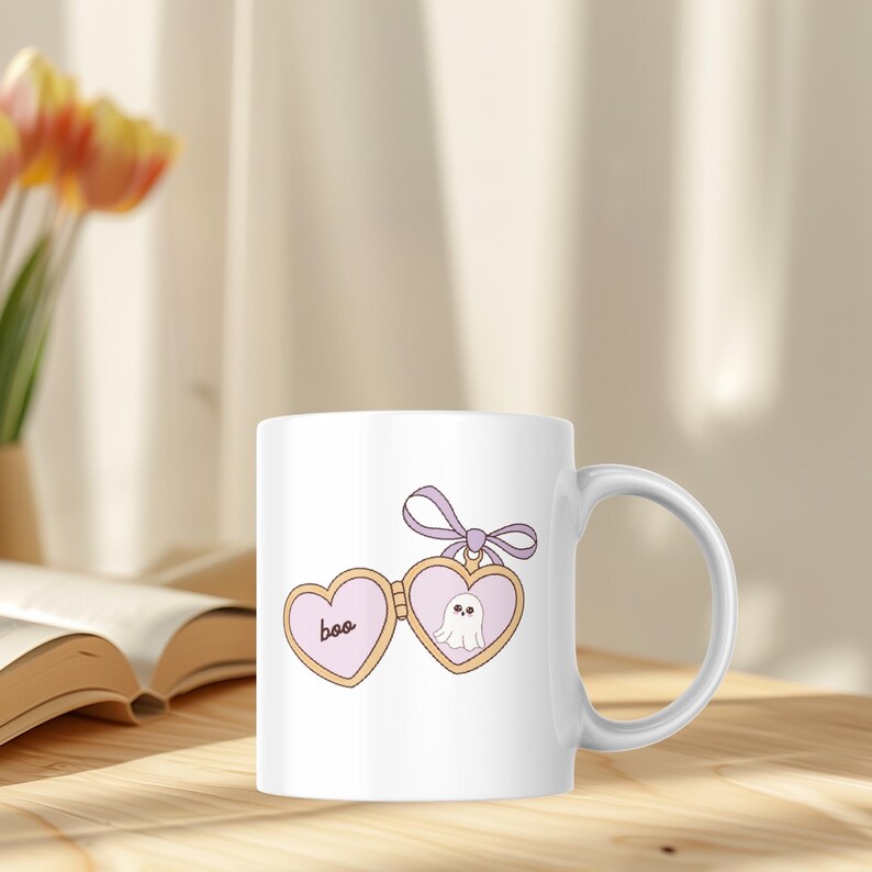 K&ouml;nnte beinhalten: Wei&szlig;e Keramiktasse mit herzf&ouml;rmigem Design, dem Wort "boo" und einer Geisterillustration. Das Design ist in Rosa-, Beige- und Lilat&ouml;nen gehalten. Die Tasse hat einen gebogenen Henkel und steht auf einer Holzoberfl&auml;che.