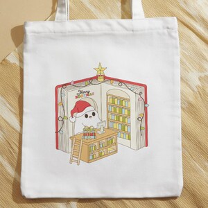 May include: White tote bag featuring a whimsical illustration of a ghost wearing a Santa hat inside a book-shaped library. The book has a red cover and the text "Merry Bookmas". The scene includes a desk, ladder, and Christmas lights.
