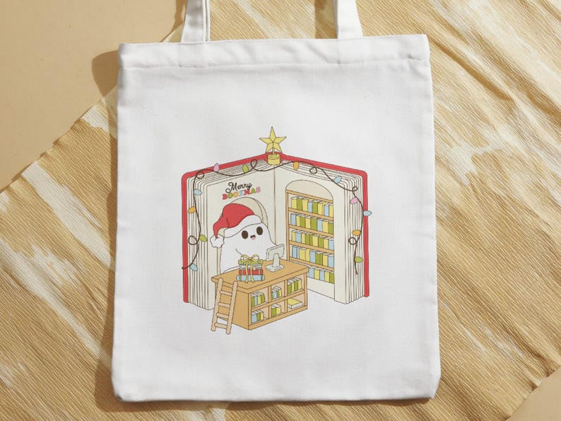 May include: White tote bag featuring a whimsical illustration of a ghost wearing a Santa hat inside a book-shaped library. The book has a red cover and the text "Merry Bookmas". The scene includes a desk, ladder, and Christmas lights.