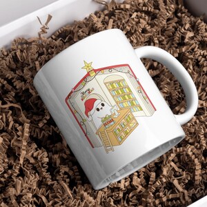May include: White ceramic mug with a Christmas-themed illustration. The design features a cartoon ghost wearing a Santa hat, inside a book with a library scene. The mug is in a white box with brown paper filler. The text "Merry Xmas" is visible.